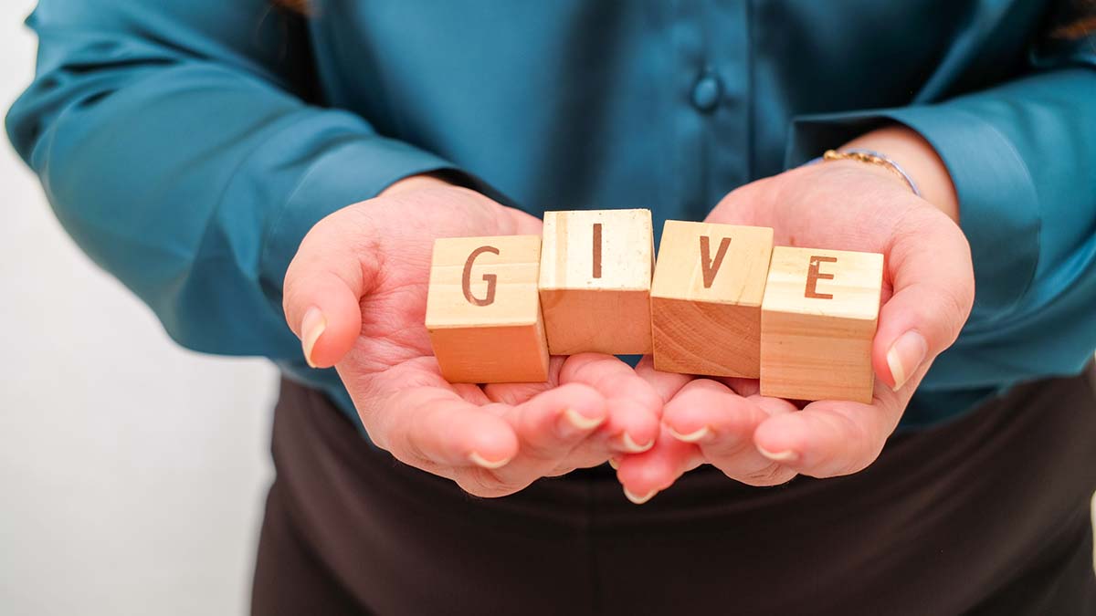 Emeritus Society | Hands hold wood blocks spelling "GIVE." Soft blue shirt frames the gesture