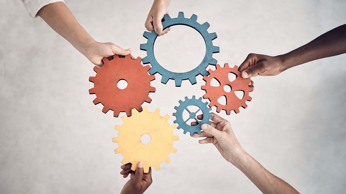 Participate | Group of five businesspeople holding and fitting gears together in an office at work. United business professionals having fun connecting gear pieces during a meeting